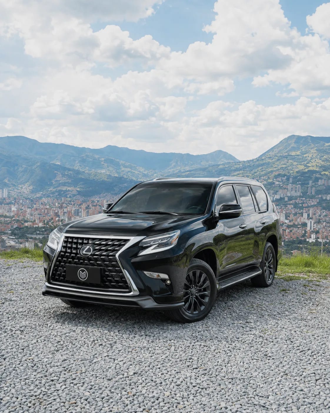 Black Lexus GX with a mesh grille, front three-quarter view, at a lookout with Medellín behind.