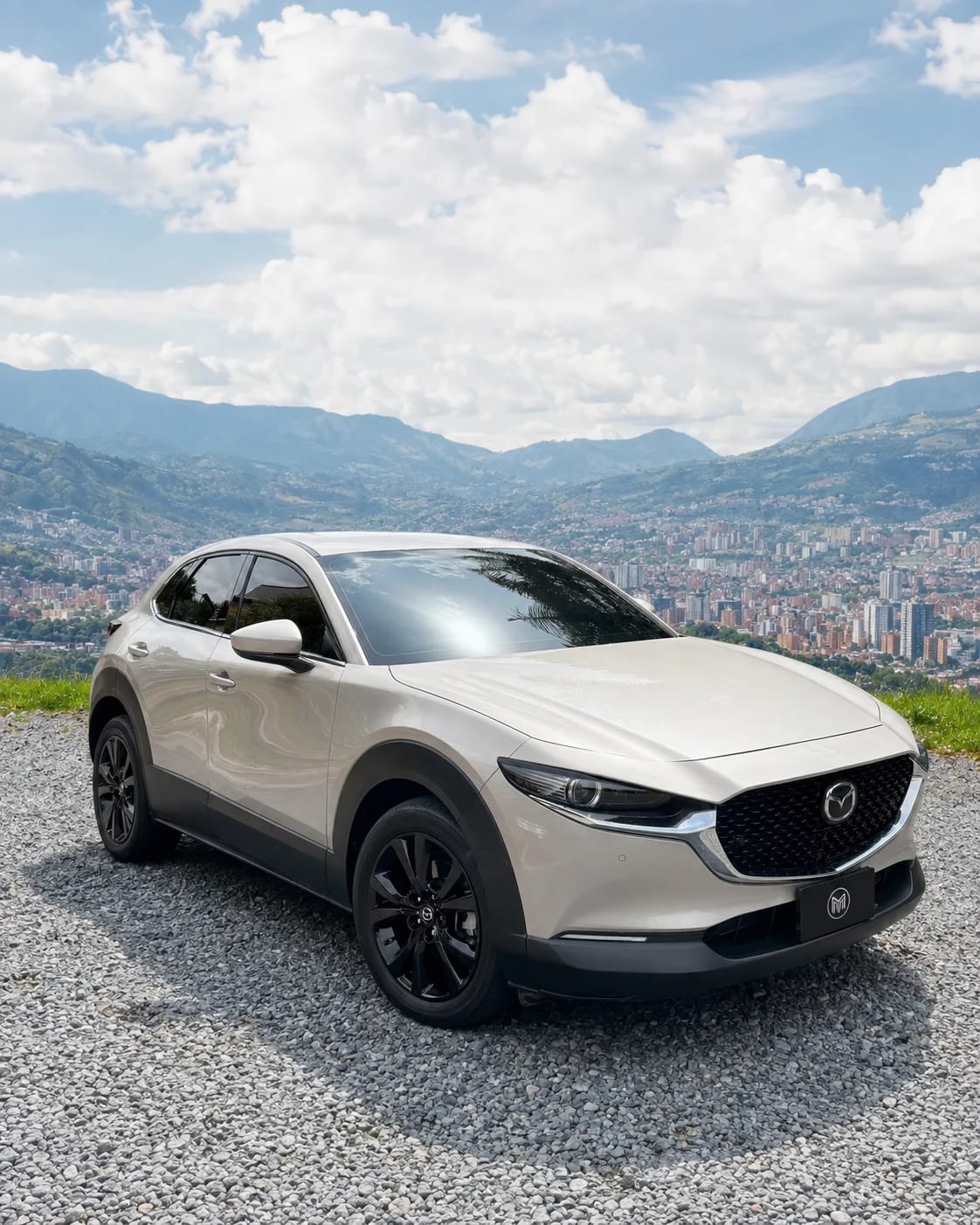 Sand white Mazda CX-30 with black wheels, front three-quarter view, at a lookout with Medellín behind.