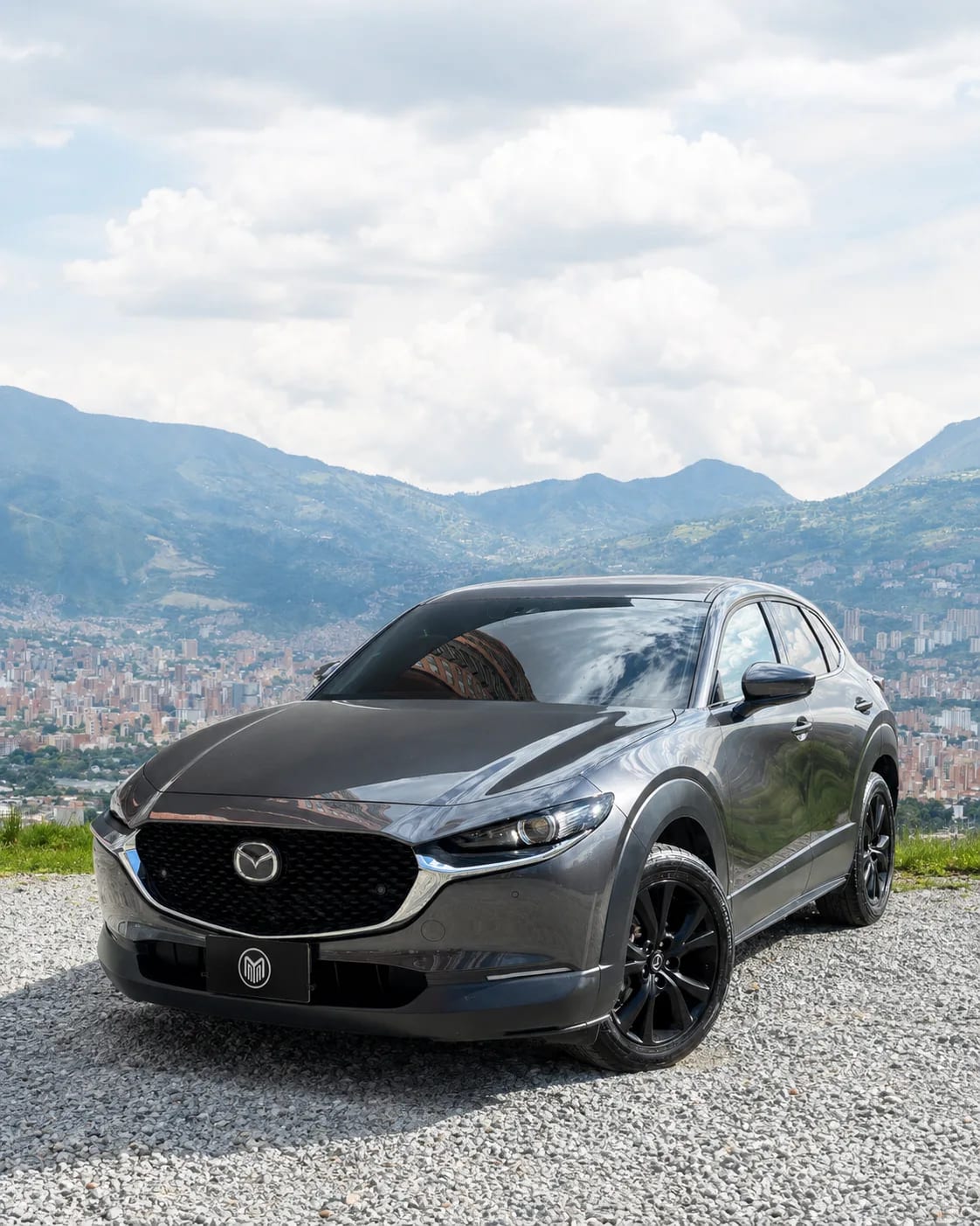 Graphite grey Mazda CX-30 with black wheels, front three-quarter view, at a lookout with Medellín behind.