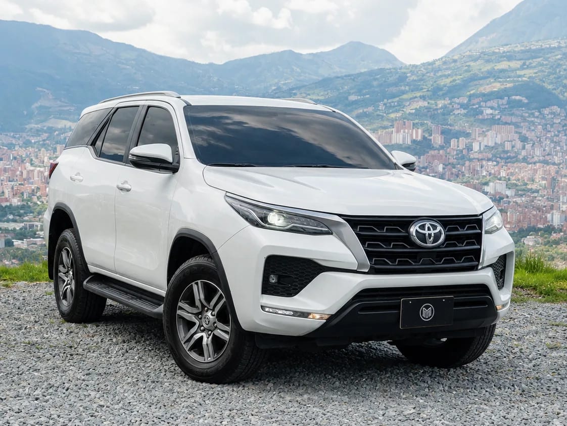 White Toyota Fortuner with roof rails, front three-quarter view, at a lookout with Medellín behind.