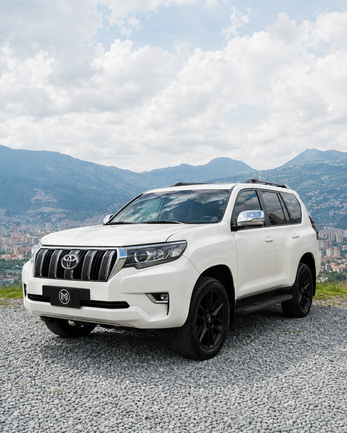 White Toyota Prado with black wheels, front three-quarter view, at a lookout with Medellín behind.