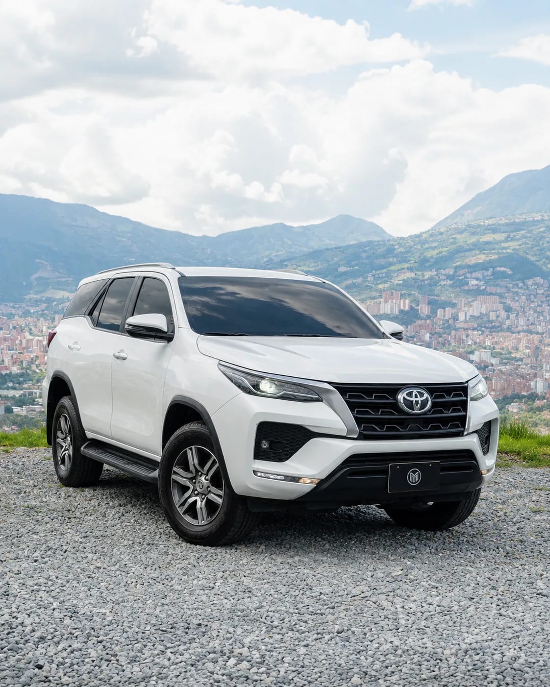 White Toyota Fortuner with roof rails, front three-quarter view, at a lookout with Medellín behind.